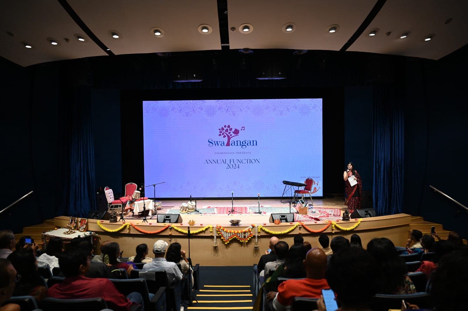 A full auditorium watching the 2024 Swarangan annual function, with a compere introducing the next item